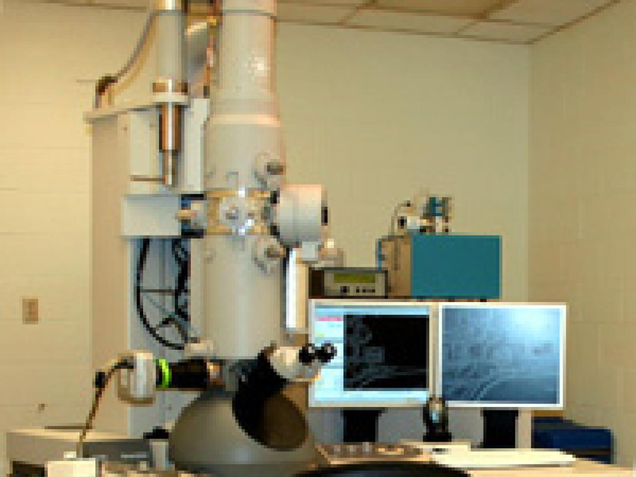 A transmission electron microscope (TEM) setup in a laboratory setting. The microscope consists of a tall vertical column in metallic silver color, with various mechanical controls and apertures visible. Two computer monitors are positioned on a desk next to the microscope for digital image viewing and instrument control. The setup appears to be in a typical research laboratory environment with ceiling tiles visible above.